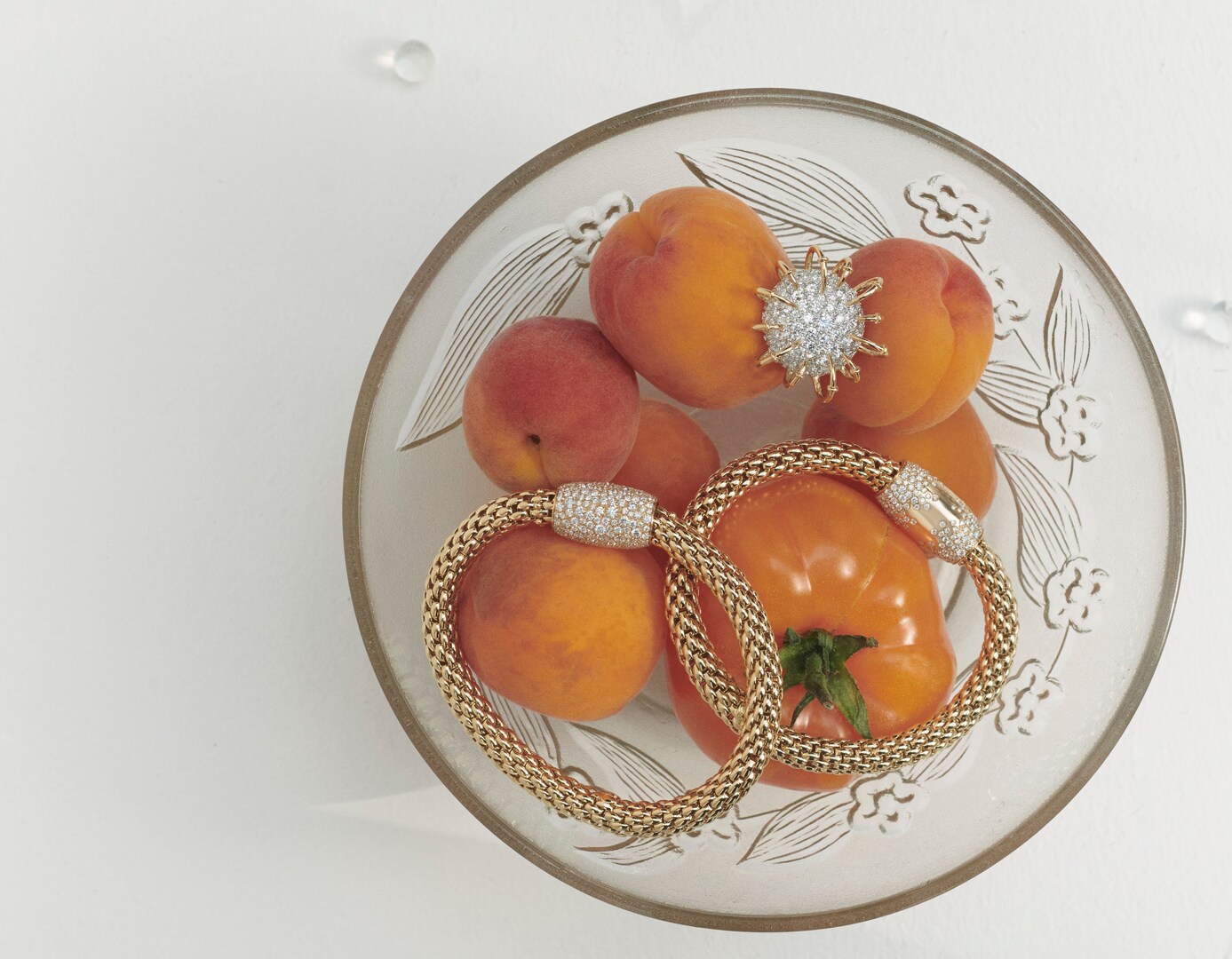 Gold bracelets and a brooch resting on apricots and a tomato in a bowl.